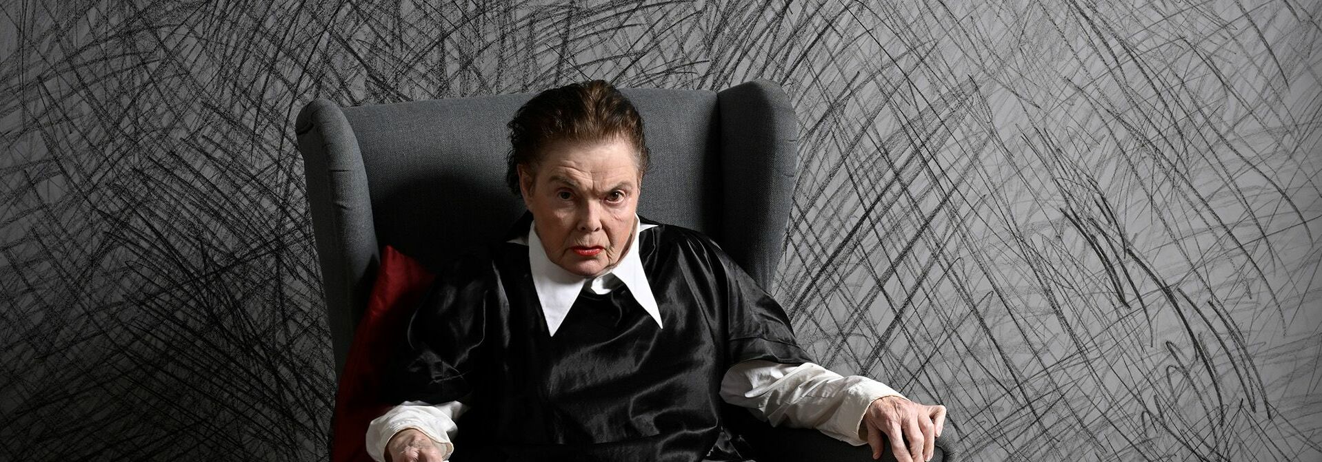 Afbeelding van de voorstelling: Een zittend persoon op een grijze stoel in nette kleding met een angstaanjagende blik. De achtergrond is een bekraste, grijze muur.