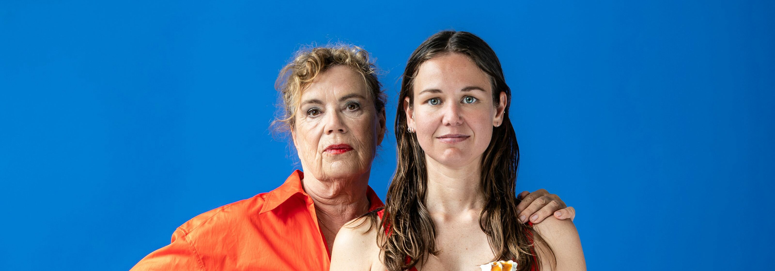 Afbeelding van de voorstelling: Twee vrouwen zijn te zien in een rode badpak, de 1 staat met een ijsje in haar hand en de ander met een strandbal. De achtergrond is blauw en ze kijken beide de lens in met een neutrale blik.