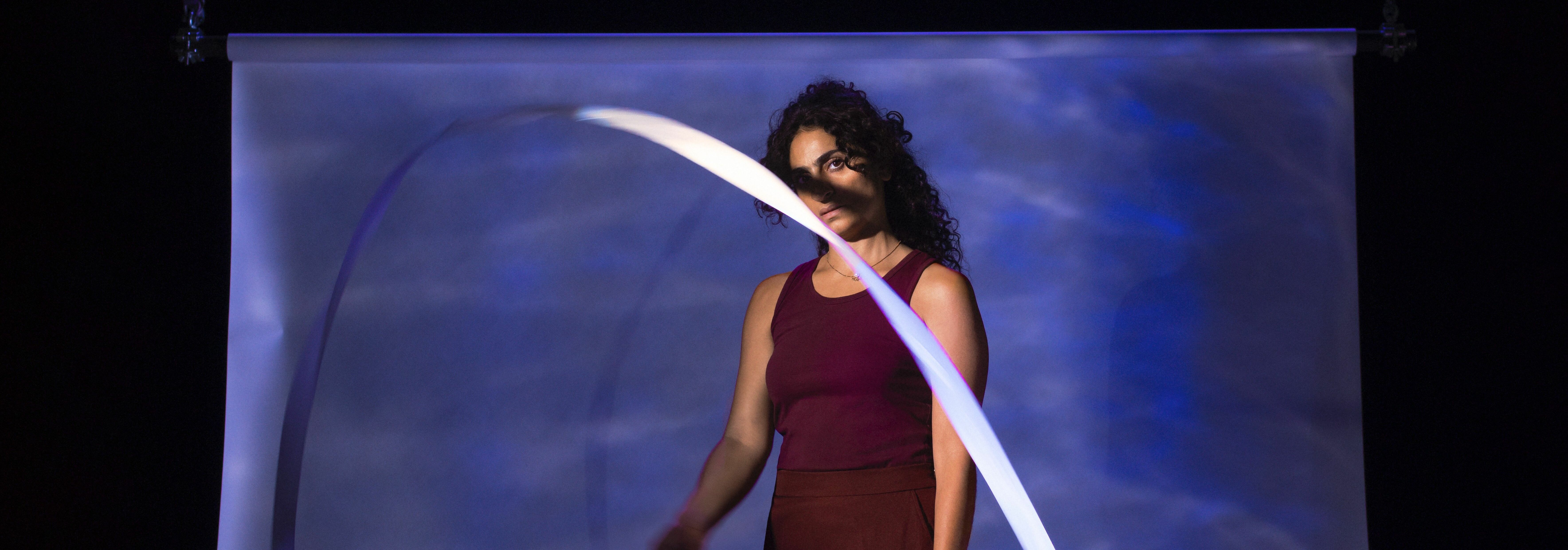 Afbeelding van de voorstelling: Een vrouw zit onderaan een trap, deze vrouw kijkt neutraal de camera in en heeft kleurrijke kleding aan met vlechten in haar haren.
