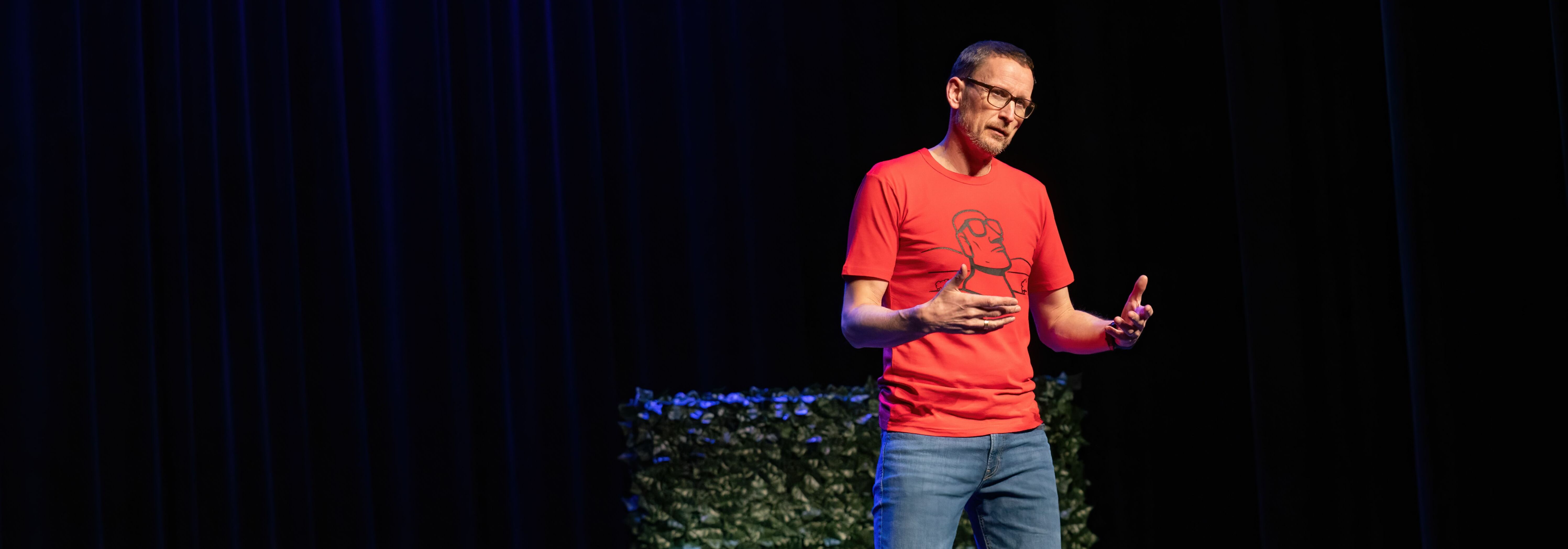 Afbeelding van de voorstelling: Een man staat met een microfoon een voorstelling te geven in nette kleding en heeft een bril op.