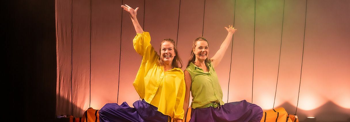 Afbeelding van de voorstelling: Een vrouw in een geel shirt en blauwe broek houdt een grote bladblazer vast en kijkt verbaasd, terwijl op de achtergrond een andere vrouw in een felgroen en oranje kostuum staat op een toneel met theaterlicht.