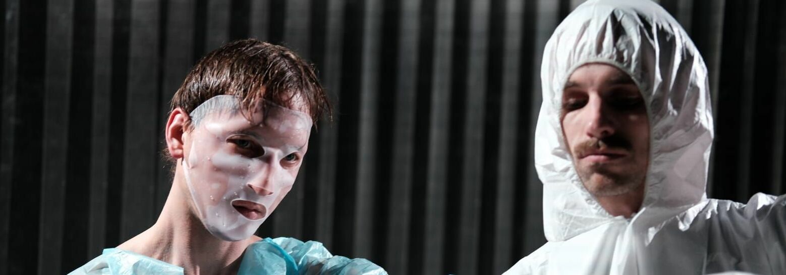 Afbeelding van de voorstelling: Een persoon in een geel pak en een wit masker staat met zijn ene hand gestrekt in een soort karate houding. De achtergrond heeft een muur met tegels en groene planten.